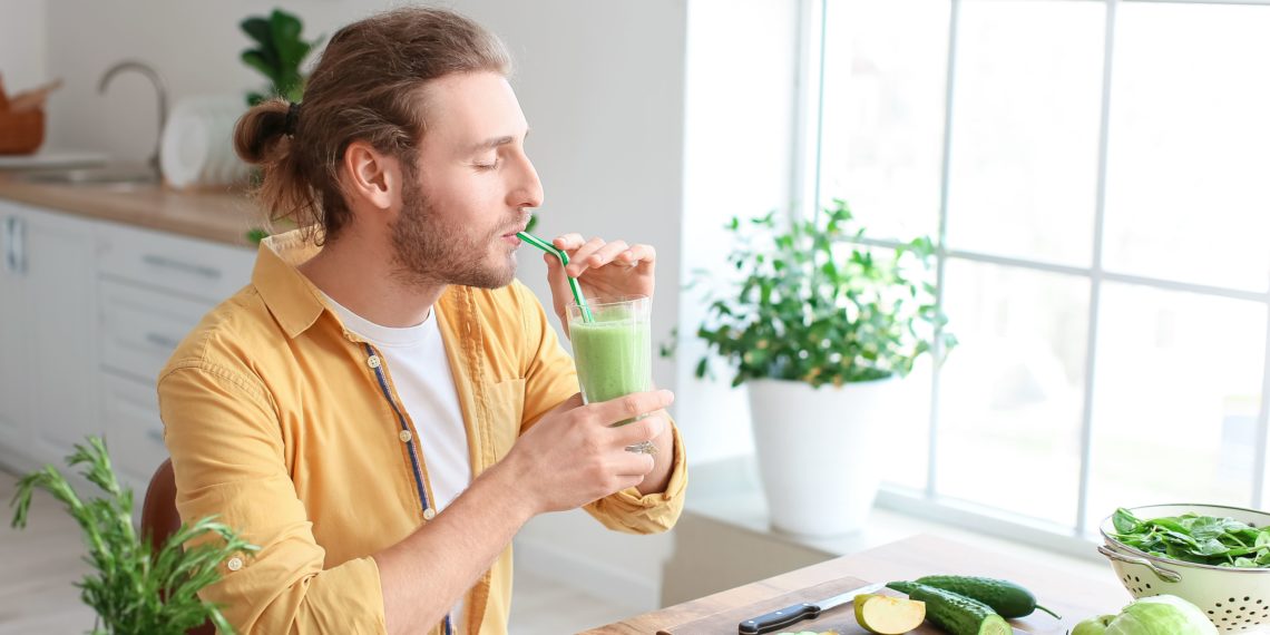 a man at his kitchen table taking a sip of a green smoothie