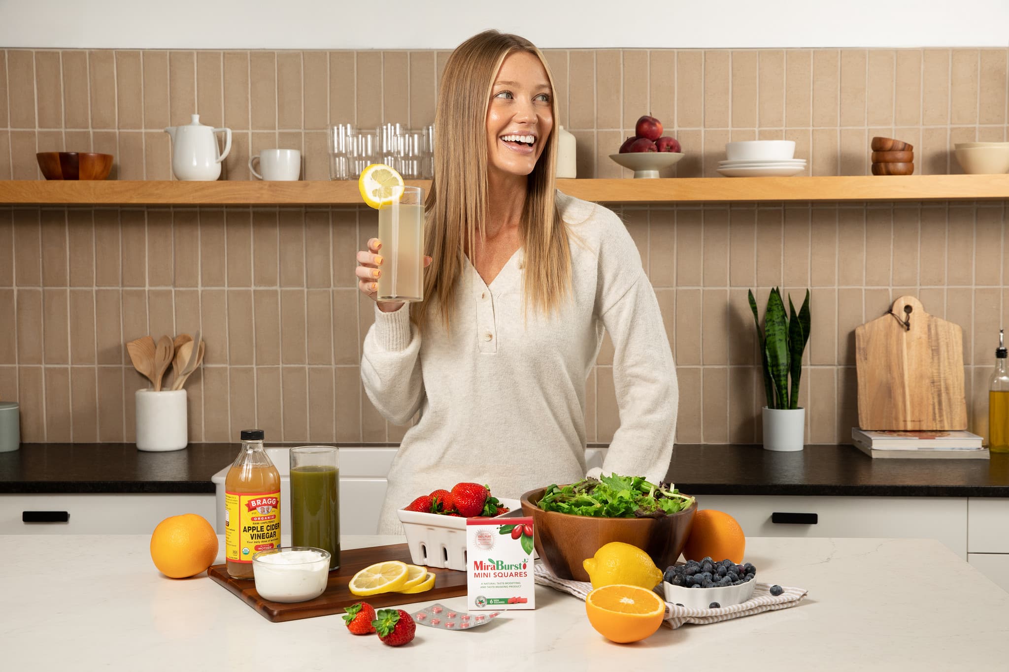 woman in a kitchen smiling with a drink in hand and MiraBurst mini squares on the counter